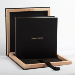 Álbum de casamento preto personalizado "Mayara e Bruno" em letras douradas, dentro de uma caixa de madeira clara aberta, com fita preta, sobre fundo branco. Ideal para noivos que buscam álbuns de fotos de casamento de luxo e fotolivros.